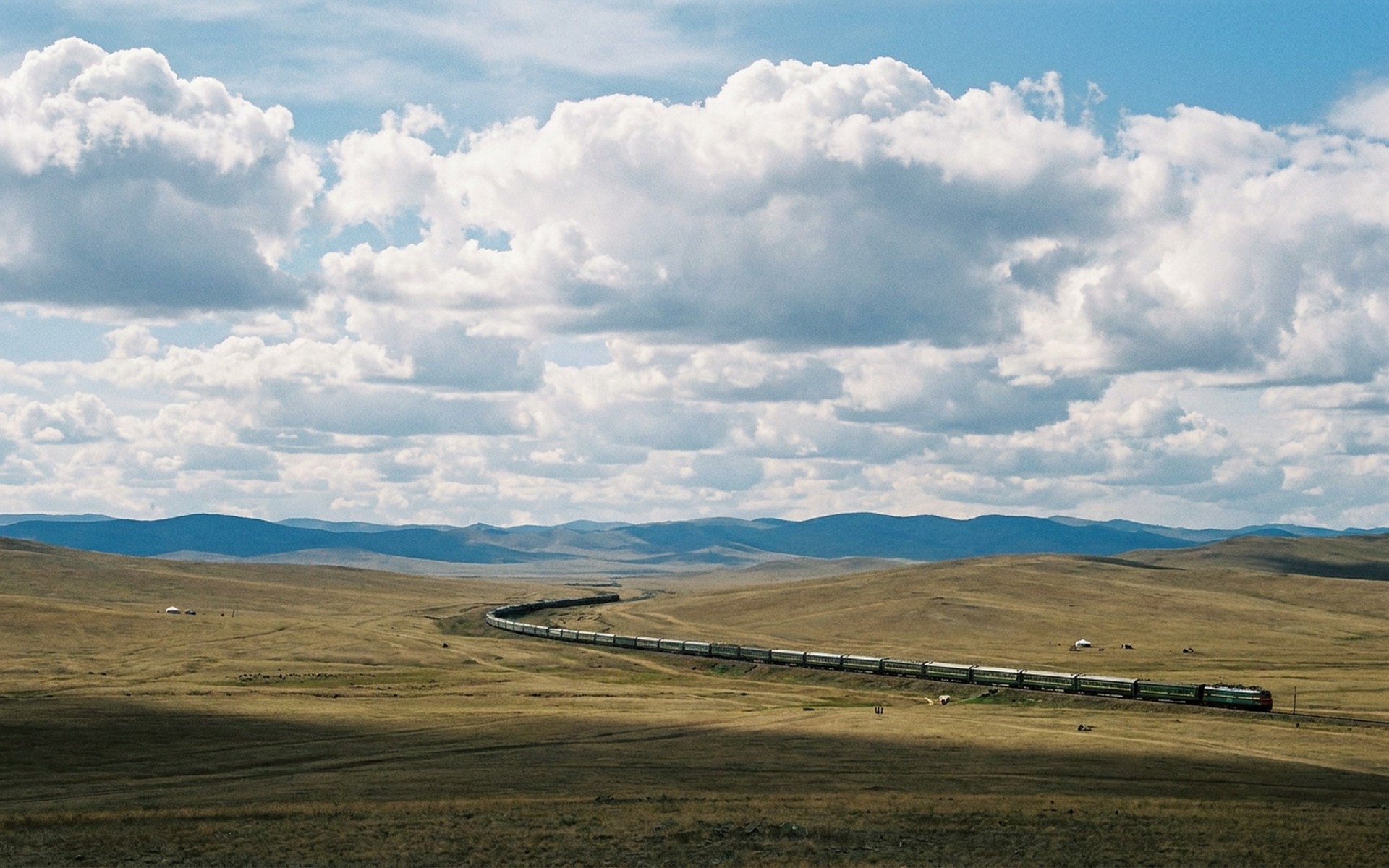 a long train curving across empty rolling steppe grassland beneath a huge sky and distant blue hills at the Trans-Mongolian route — original Triple VIP destination visual.