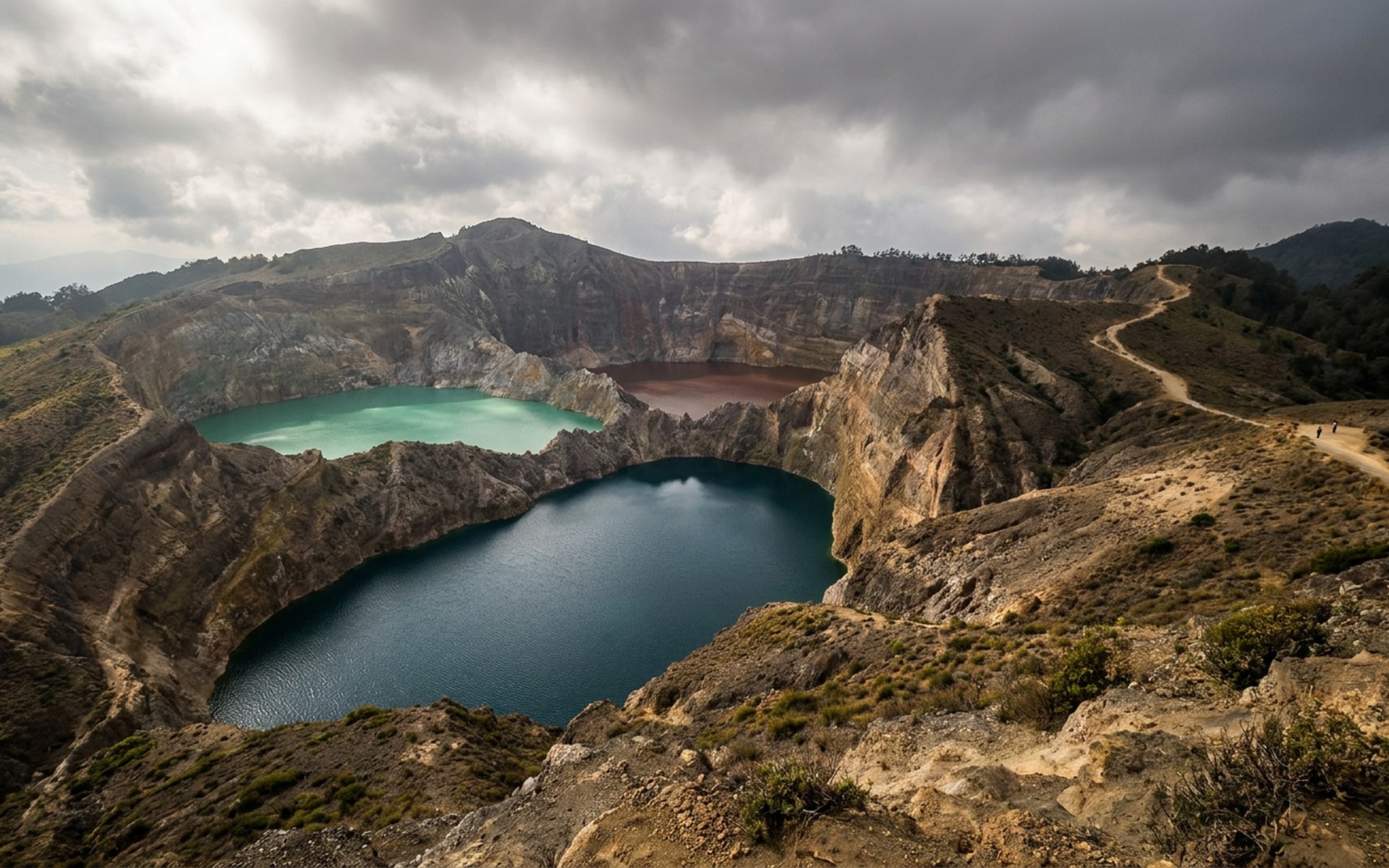 The stunning, vividly colored crater lakes of Mount Kelimutu, each a distinct hue, viewed from above under a dramatic cloudy sky, dry season. at Kelimutu & Central Flores, Indonesia — original Triple VIP destination visual.