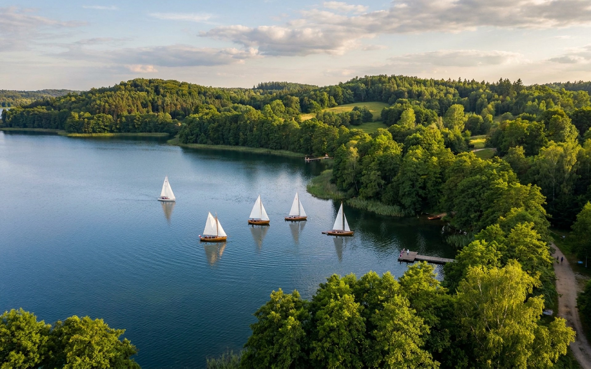 Sailboats gliding on a serene, deep blue lake, surrounded by dense green forests and rolling hills under a gentle summer afternoon sky. at Masurian Lakes, Poland — original Triple VIP destination visual.