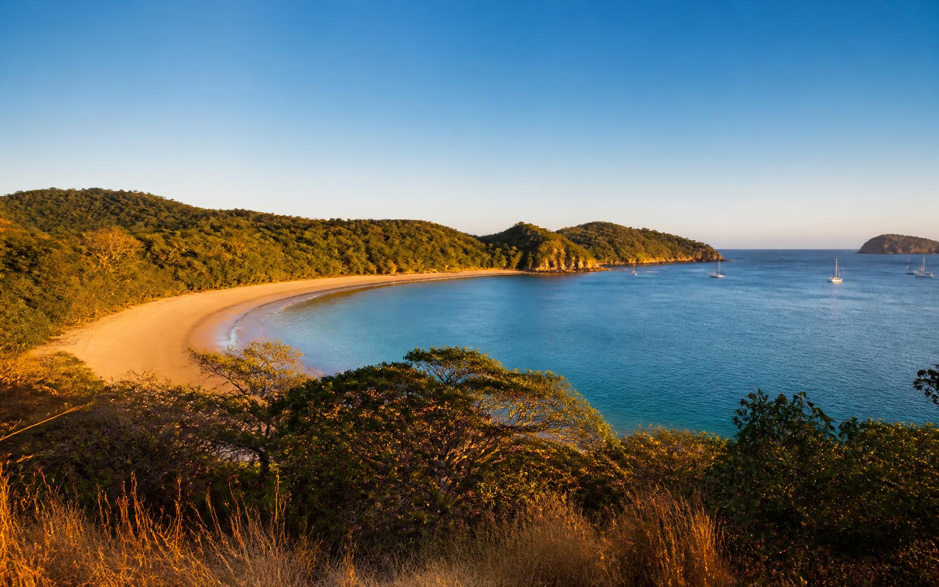 a crescent of pale gold beach curving around a calm blue-green Pacific bay on the Guanacaste coast of Costa Rica at golden hour, with dry tropical forest headlands on either side and sailboats anchored offshore - original Triple VIP destination visual.