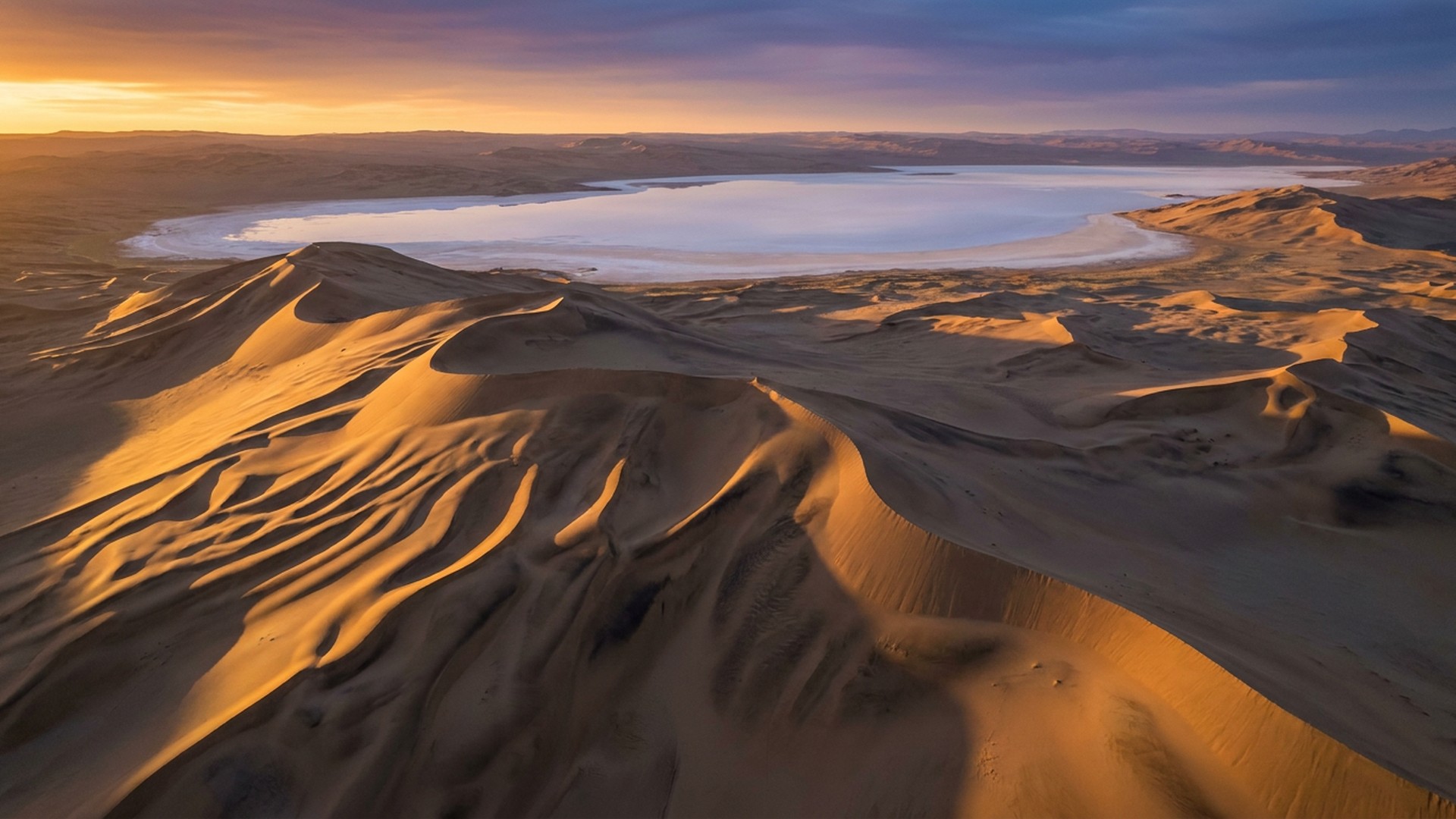 Wind-carved desert dunes and a distant salt pan at dawn