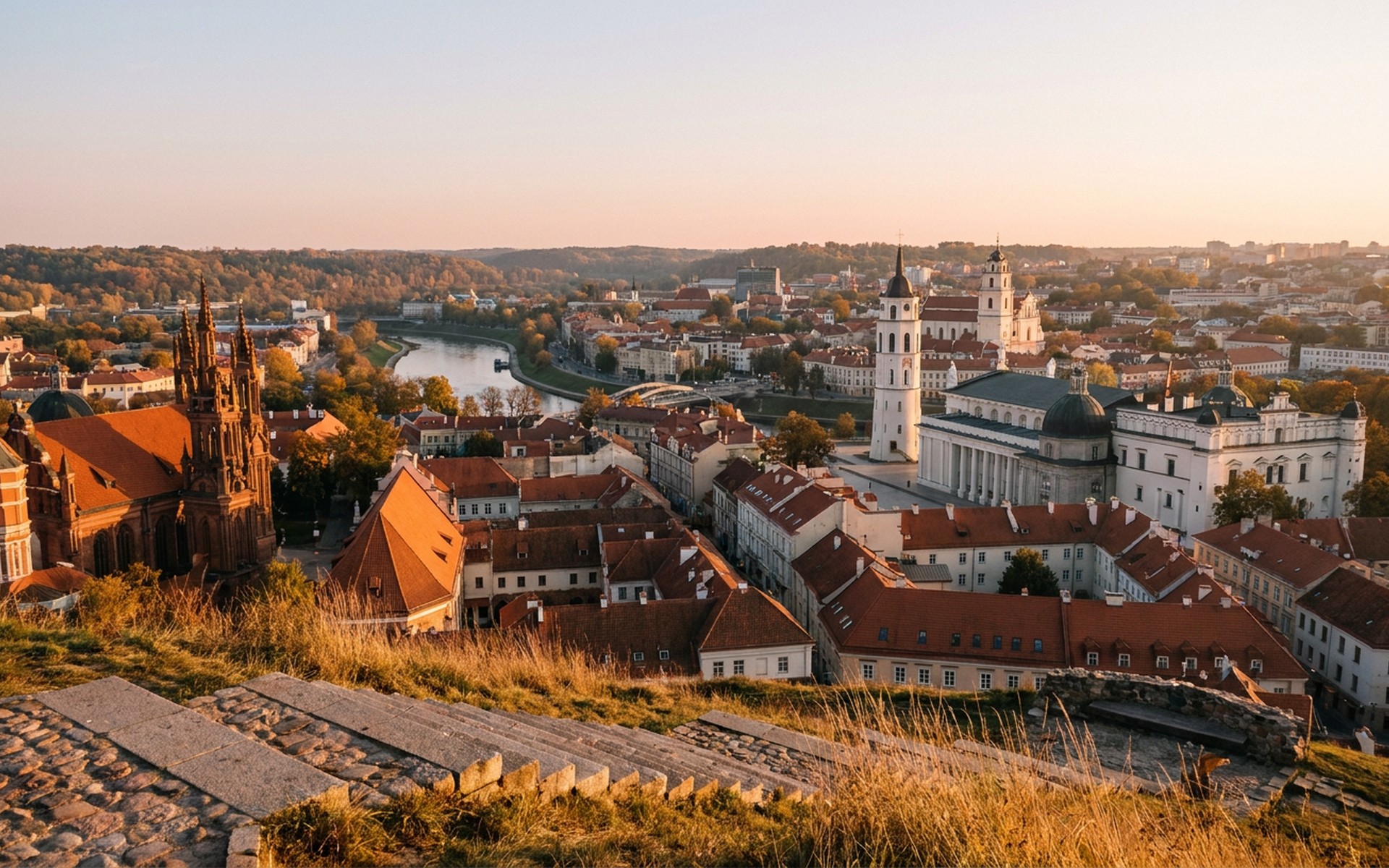 baroque church spires and red-tiled old-town roofs seen from a hill above the city at golden hour at Vilnius, Lithuania — original Triple VIP destination visual.
