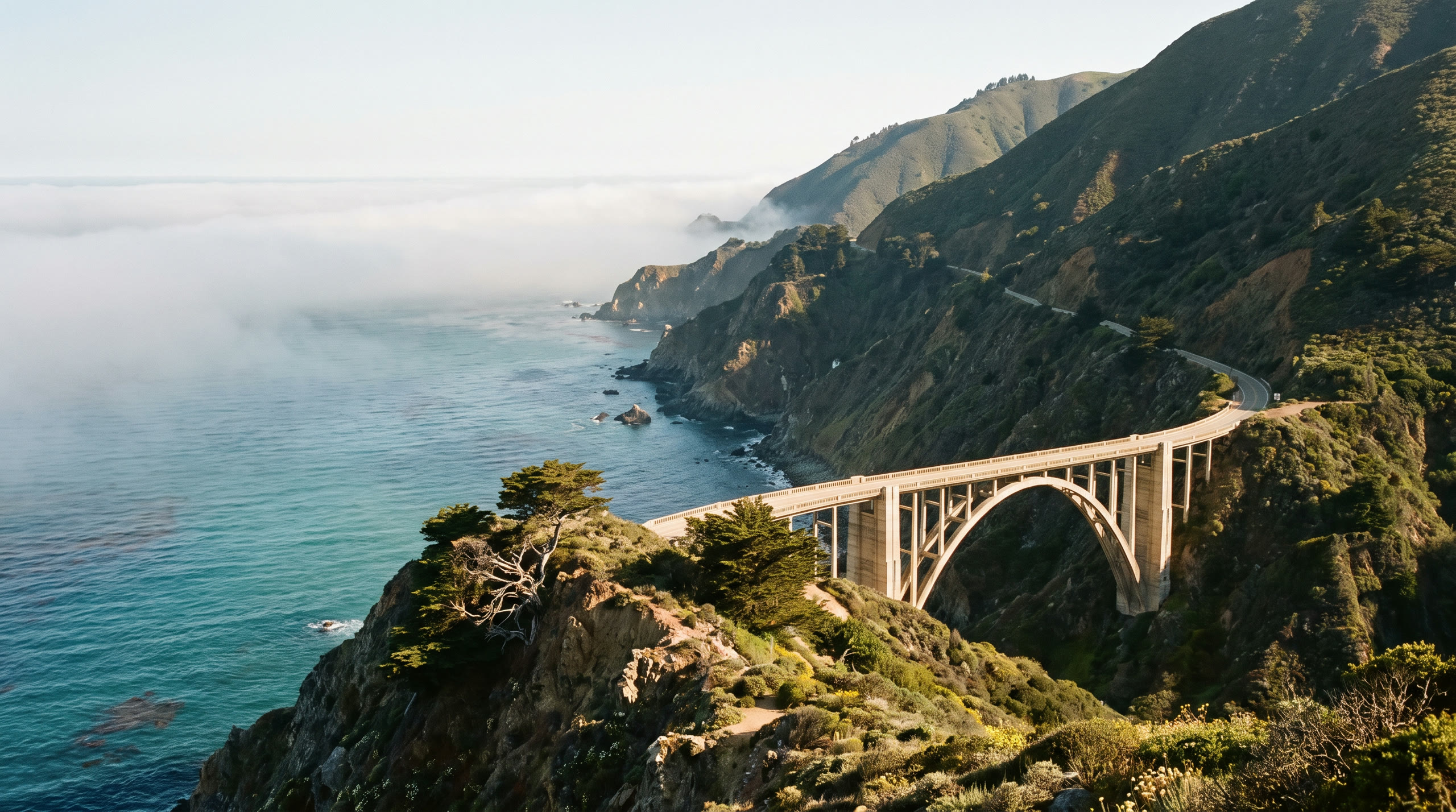 the coast highway bridging a ravine above the Pacific cliffs at Big Sur Coast, United States, in late afternoon light — original Triple VIP destination visual.