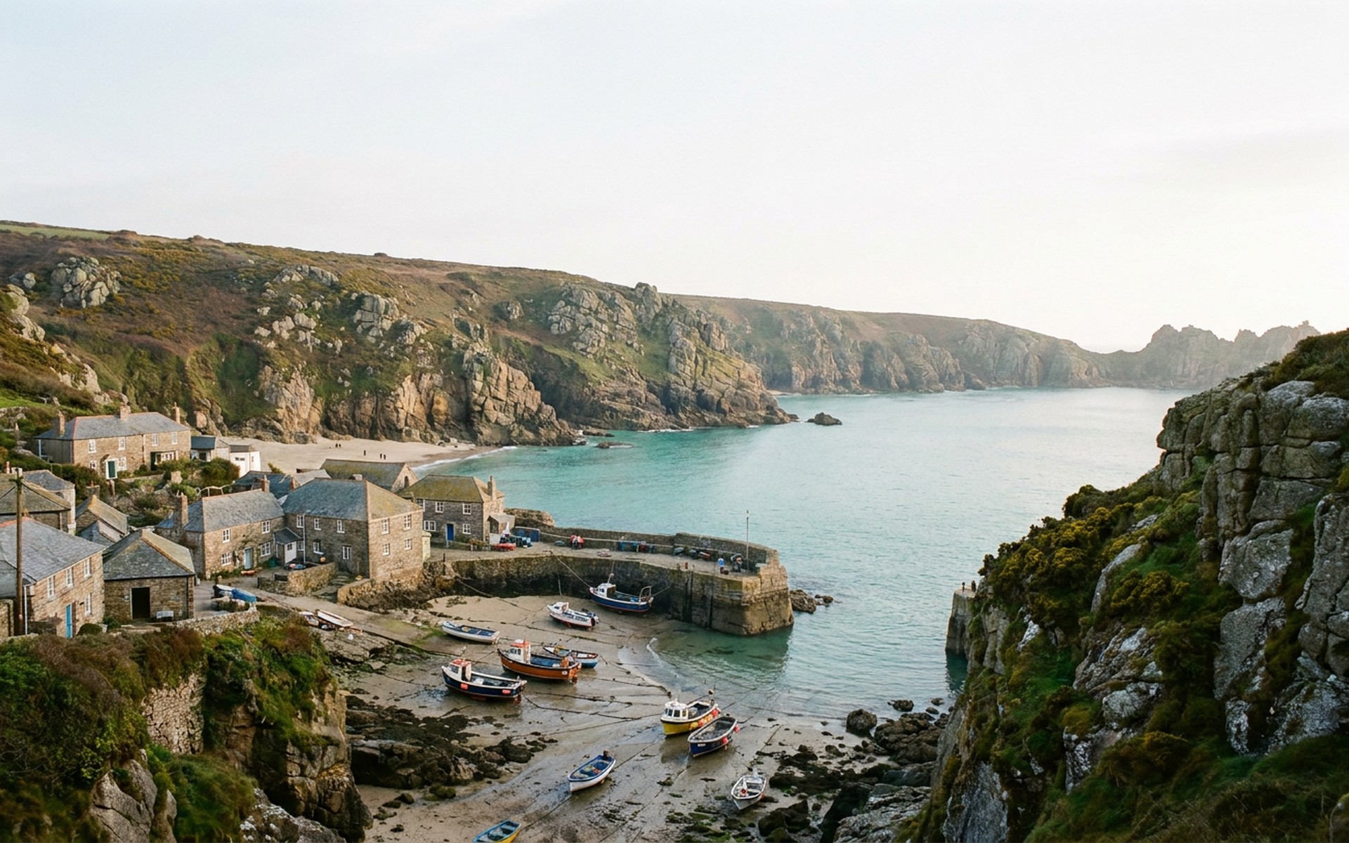 a turquoise cove below granite cliffs with a small harbour village and fishing boats at low tide at Cornwall, England — original Triple VIP destination visual.