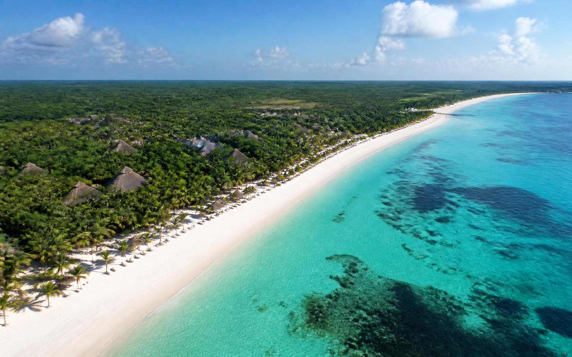 an aerial view of a long white Caribbean shore with turquoise shallows, dark offshore reef and palapa-roofed resorts set back in the jungle at Riviera Maya, Mexico, in mid-morning light — original Triple VIP destination visual.