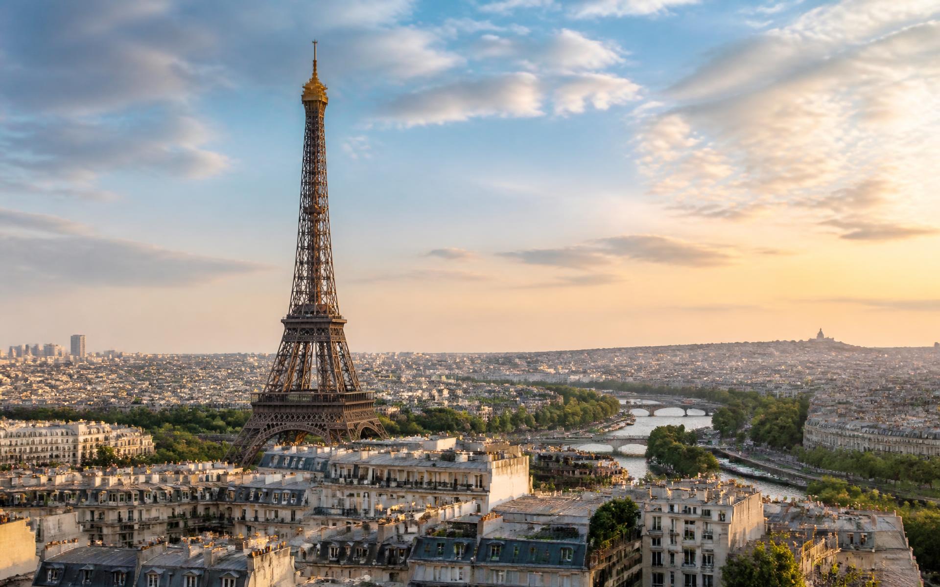 Wrought-iron Paris tower rising above Haussmannian zinc rooftops with the Seine curving through the city at golden hour