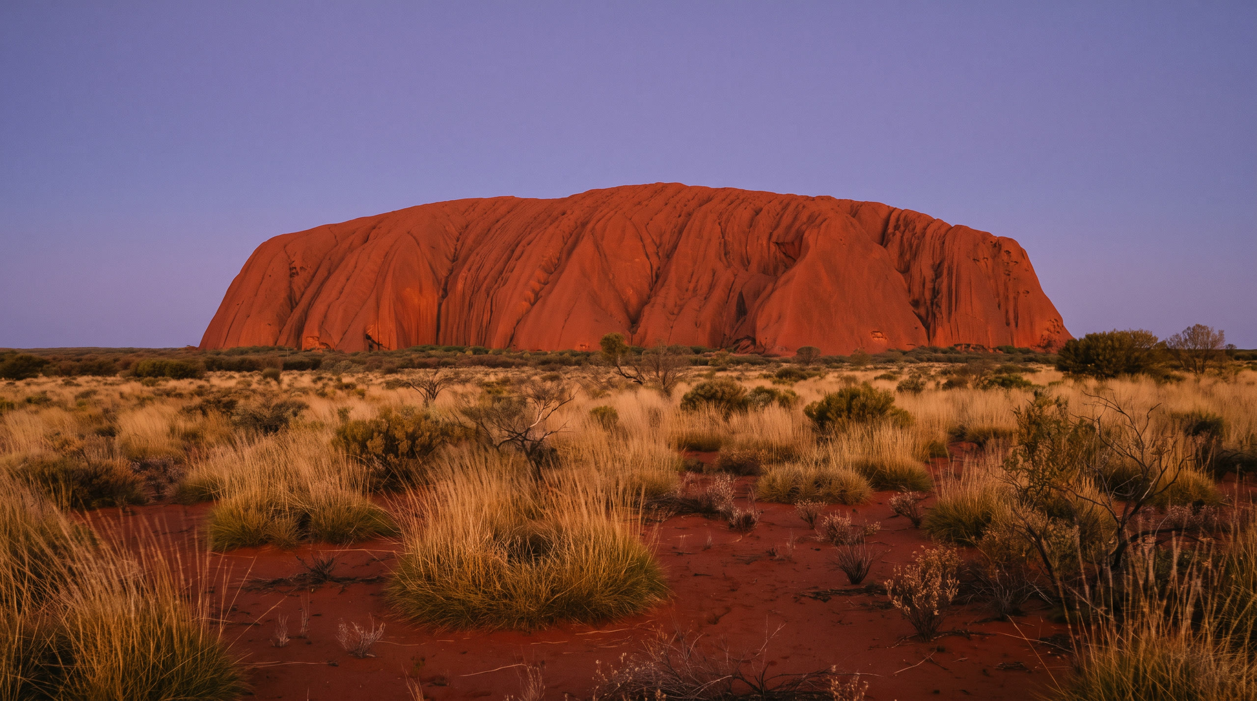 the monolith glowing deep red above the spinifex plain at Uluru & the Red Centre, Australia, in sunset light — original Triple VIP destination visual.