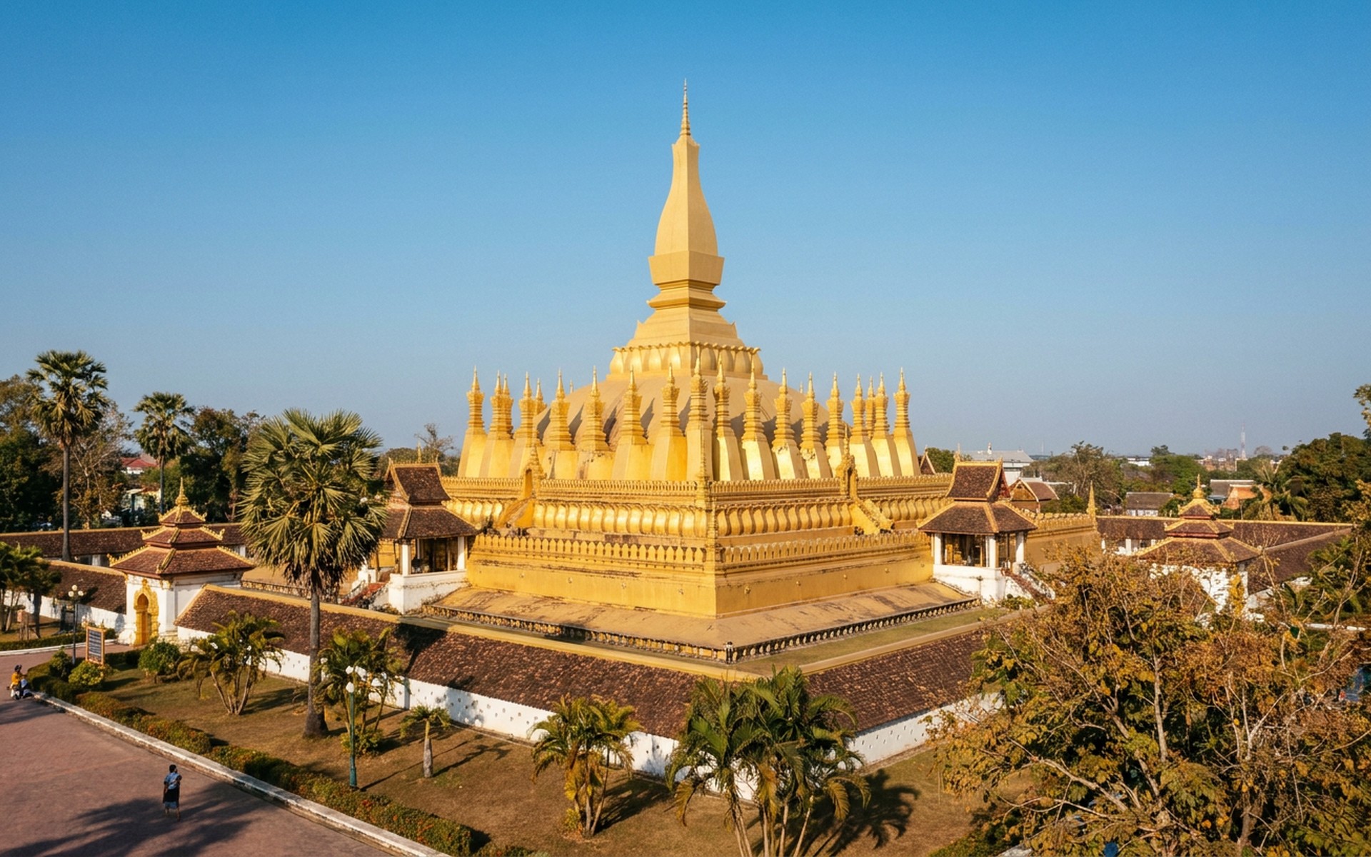 Golden stupa of Pha That Luang under a clear blue sky, with tropical trees surrounding the complex during the dry season. at Vientiane, Laos — original Triple VIP destination visual.