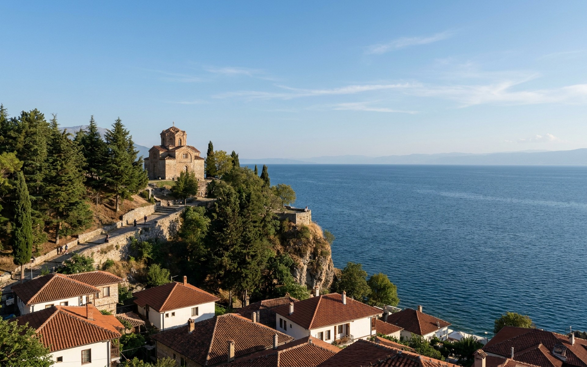 a stone church on a wooded bluff above a deep blue lake with red-roofed old town below at Lake Ohrid, North Macedonia — original Triple VIP destination visual.