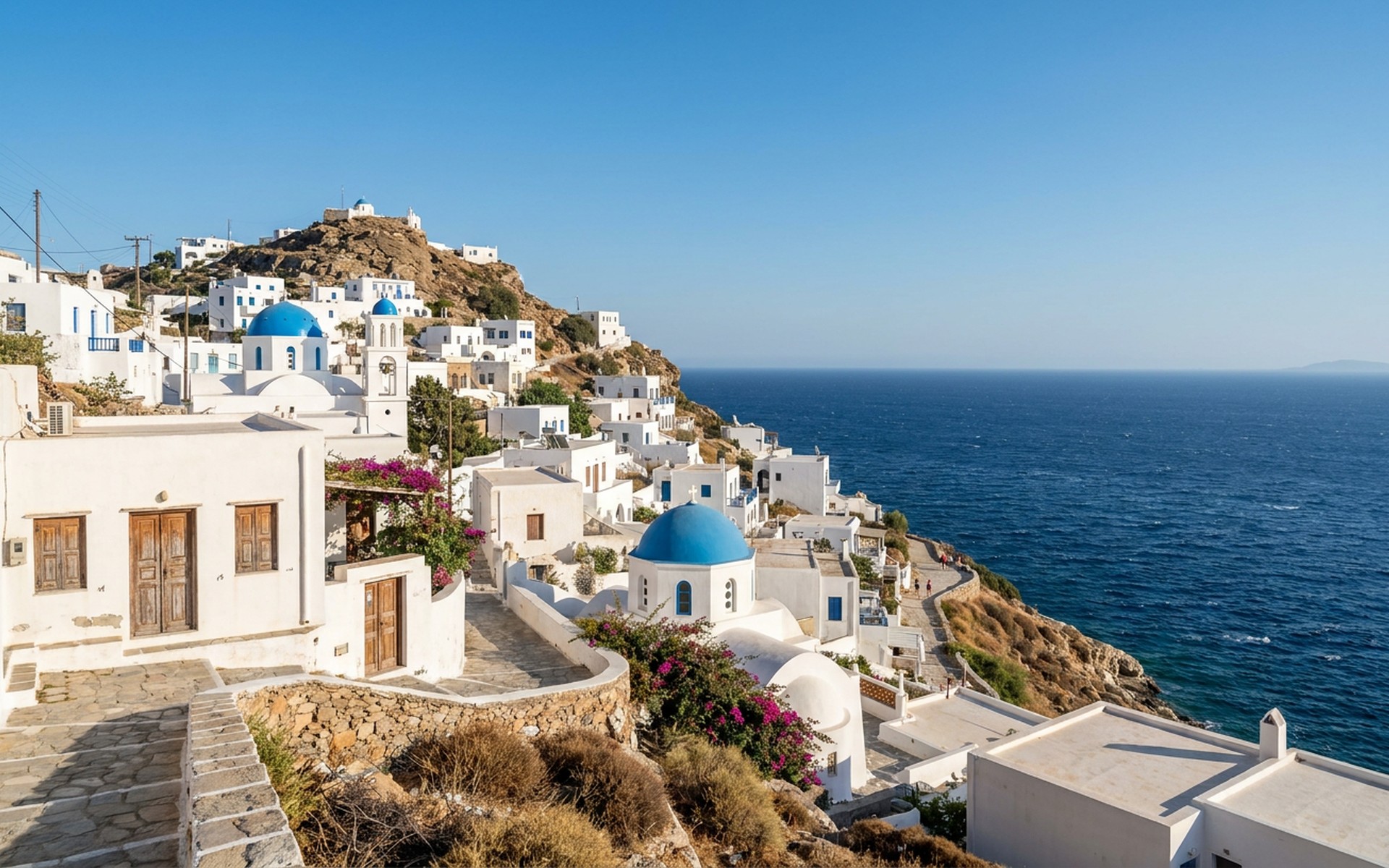 Whitewashed Cycladic architecture with blue domed churches cascading down a hillside towards a sparkling Aegean Sea, under a clear summer sky. at Ios, Greece — original Triple VIP destination visual.