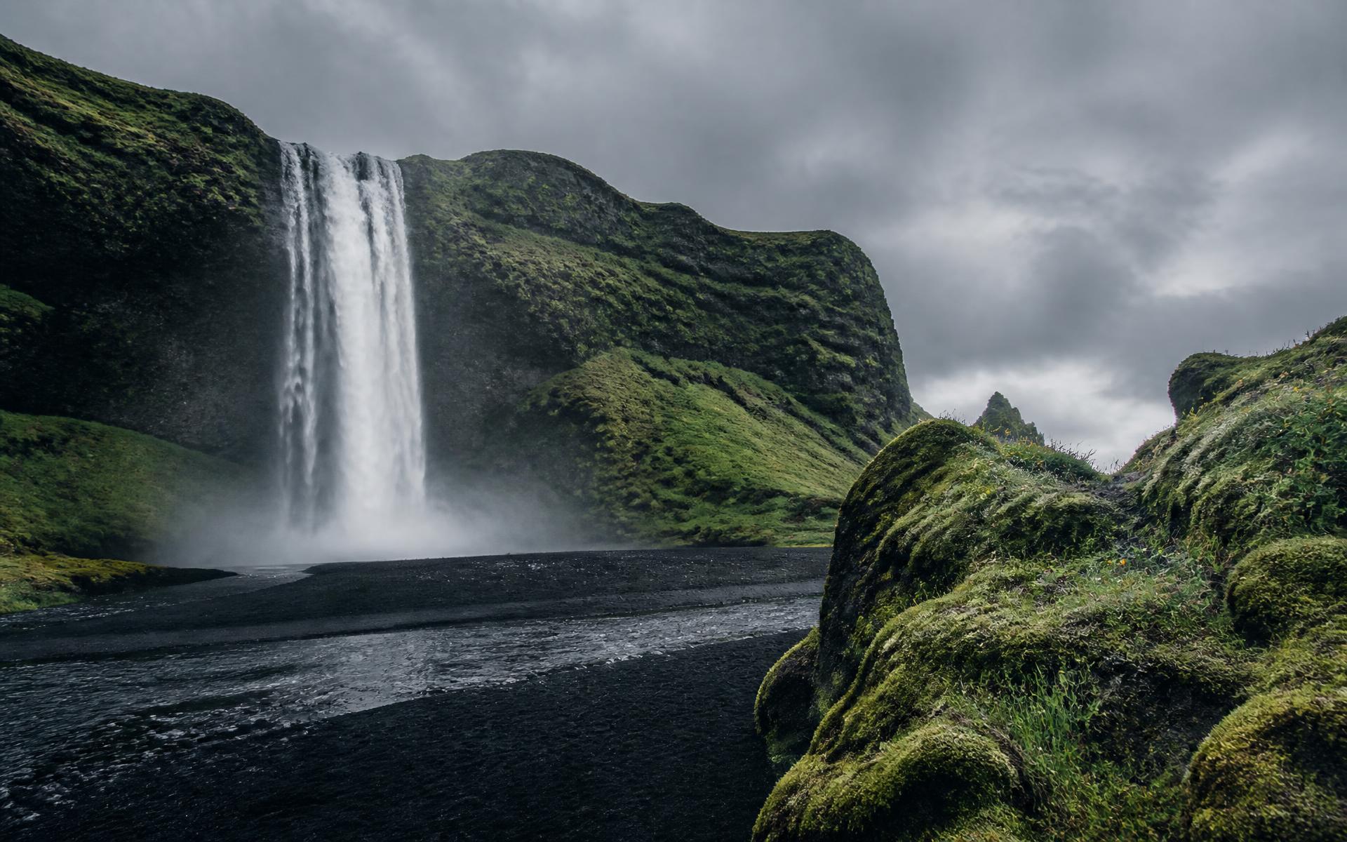 A powerful waterfall falling over moss-covered cliffs onto black volcanic sand in Iceland