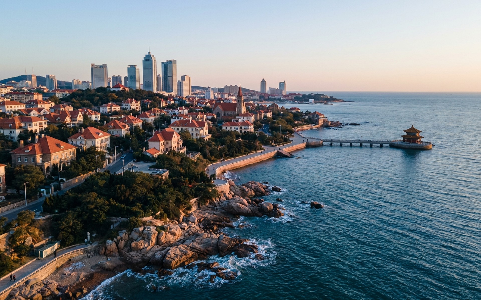 Qingdao at golden hour with red-roofed historic quarter above the rocky Yellow Sea shore, pier pavilion and modern skyline beyond