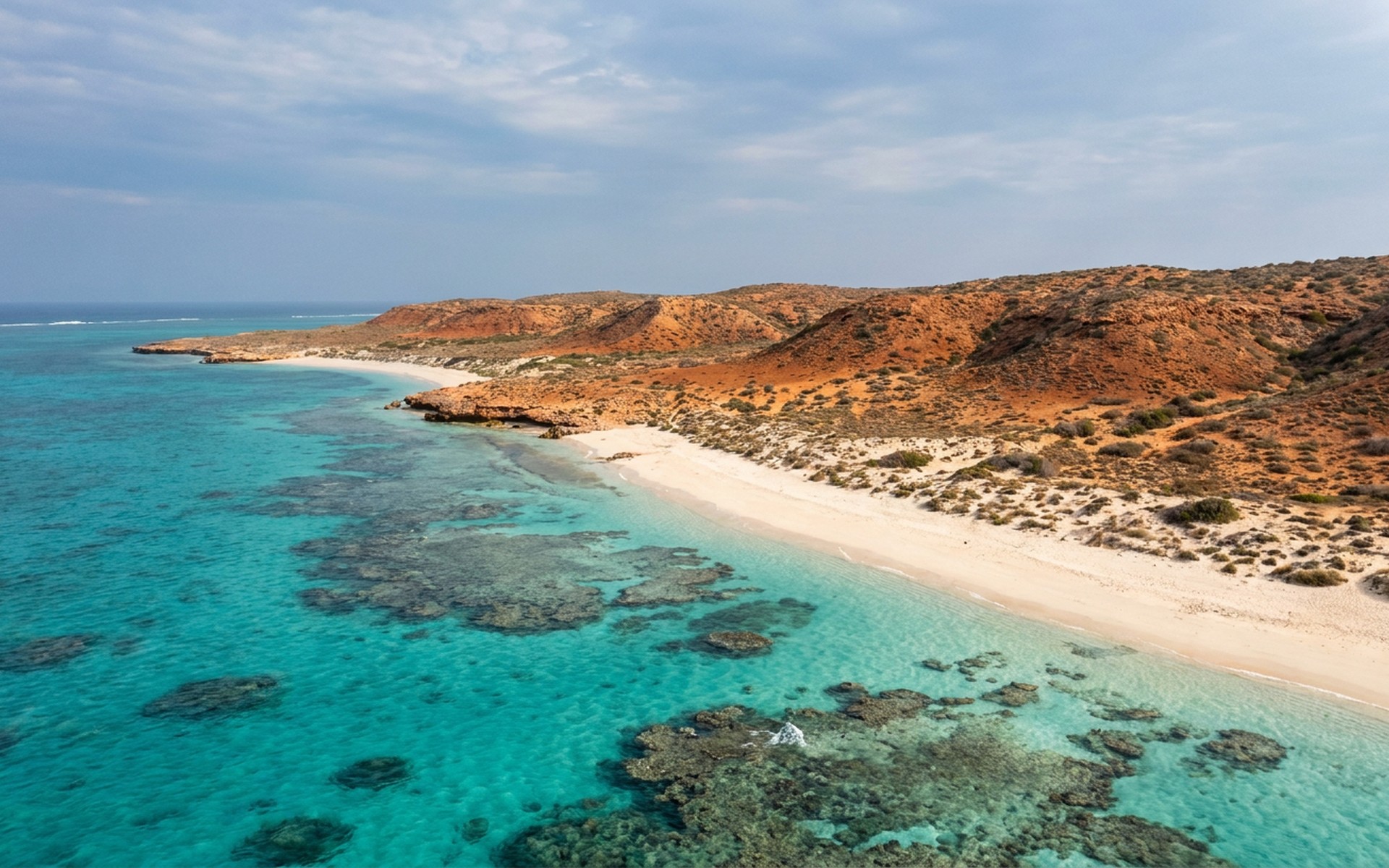 a fringing coral reef in clear turquoise water right against a red arid coastline of low ridges and white sand at Ningaloo Reef, Australia — original Triple VIP destination visual.