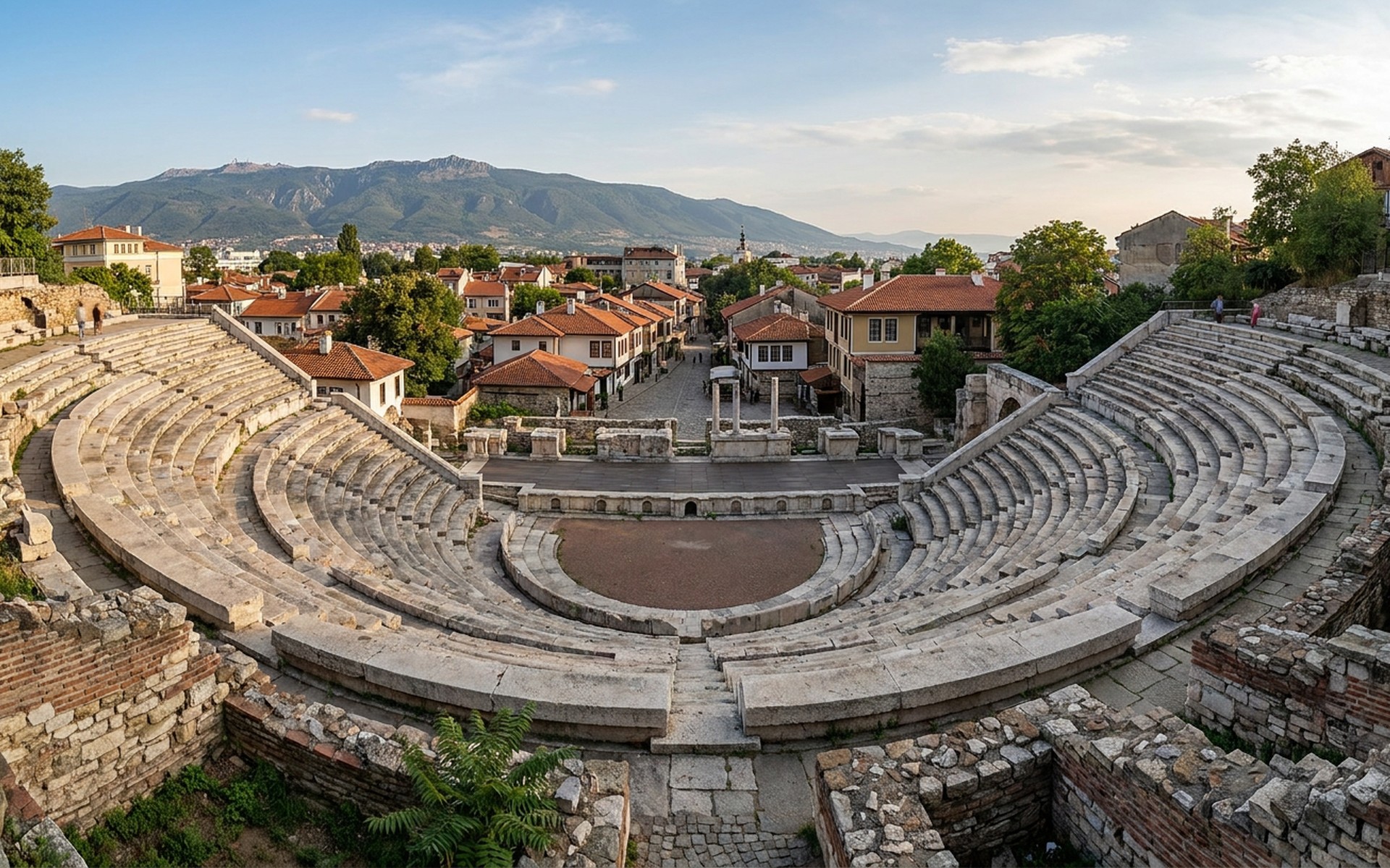 a Roman amphitheatre of tiered stone seats above red-roofed houses and a distant mountain ridge at Plovdiv, Bulgaria — original Triple VIP destination visual.