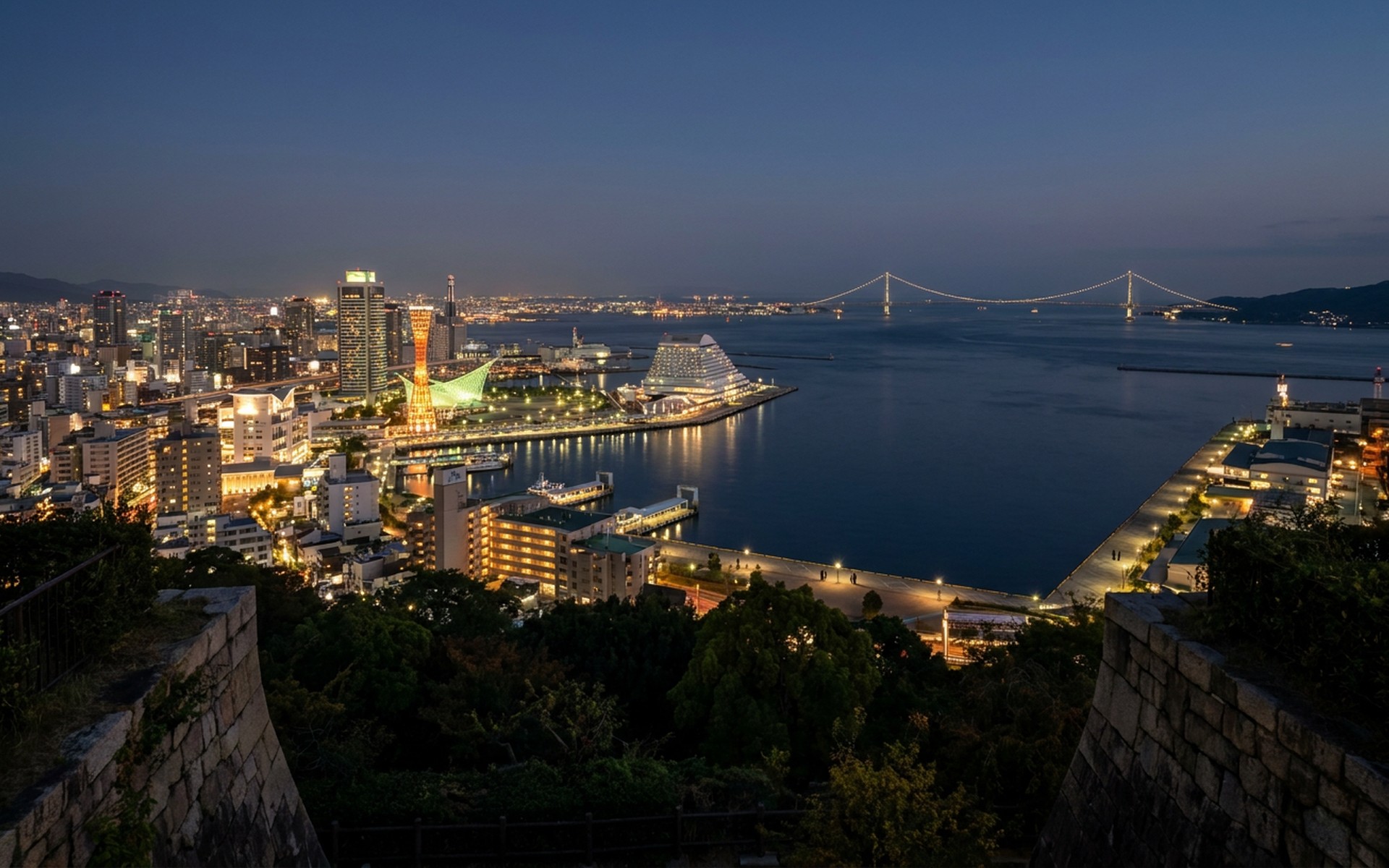 Panoramic night view of Kobe's illuminated cityscape and port, with Akashi Kaikyō Bridge visible in the distance over the calm Inland Sea. at Kobe & the Inland Sea, Japan — original Triple VIP destination visual.
