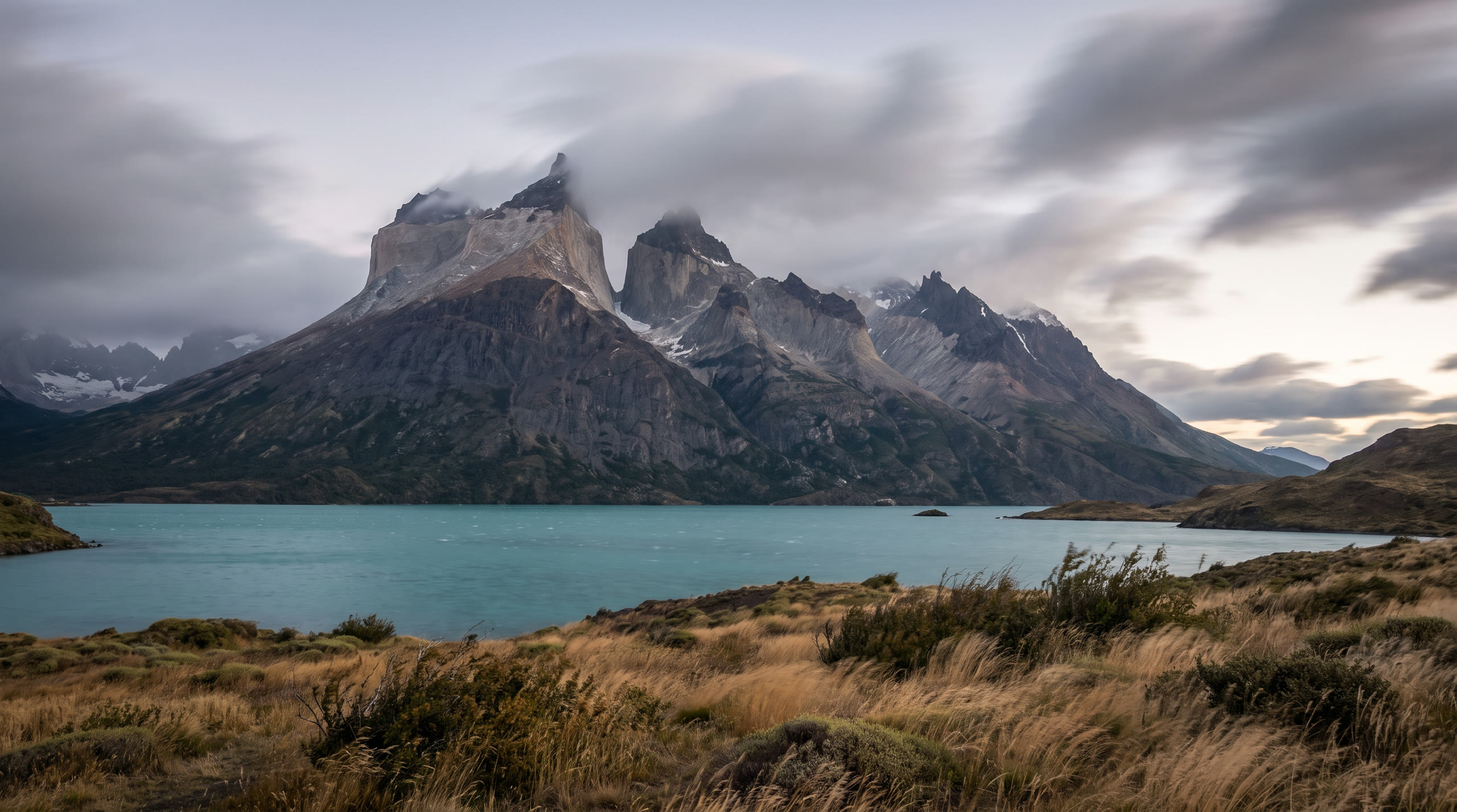 the granite towers above a turquoise glacial lake and wind-bent grassland at Torres del Paine, Chile, in first light light — original Triple VIP destination visual.
