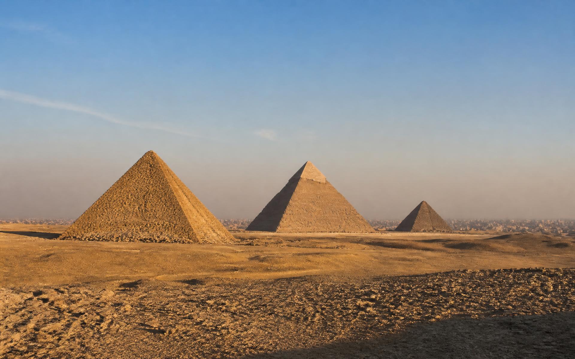 The three Giza pyramids in diagonal alignment across the desert plateau with Cairo faint on the horizon in late afternoon light